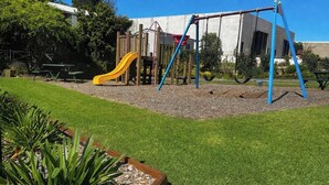 Un parque infantil con un tobogán amarillo y columpios, rodeado de césped y un edificio al fondo.