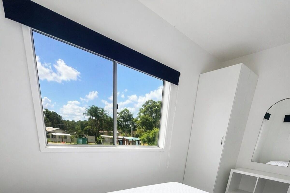 Habitación de hotel con una cama grande, un armario, una mesita de noche y una ventana con vistas a zonas verdes y edificios.