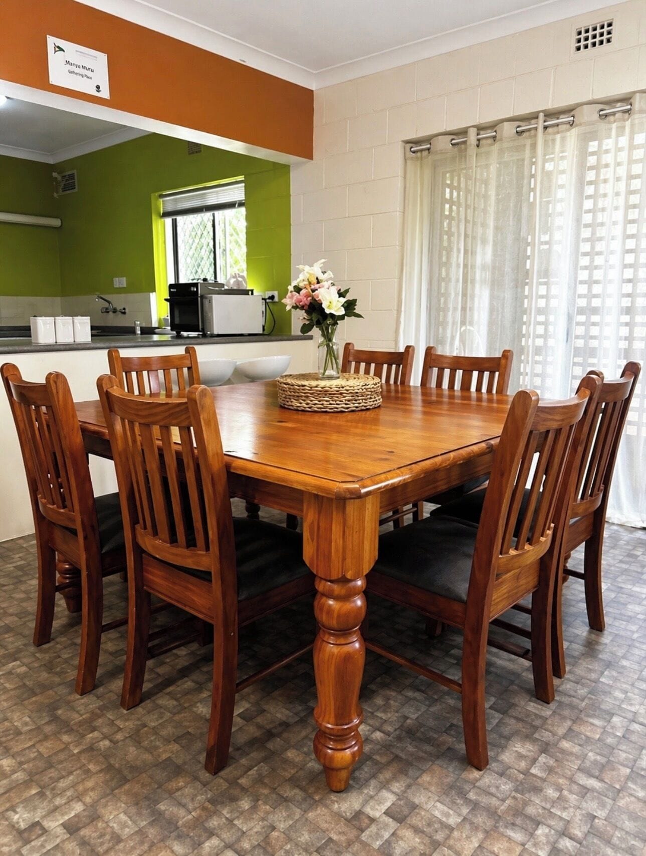 A dining area with a wooden table and chairs, a vase with flowers, and a kitchen in the background.