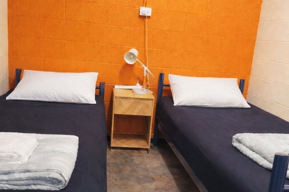 Una habitación pequeña y funcional con dos camas individuales, una mesita de noche con lámpara y un enchufe en la pared.