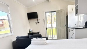 Una habitación de hotel moderna con una cama grande, un televisor de pantalla plana en la pared, una kitchenette pequeña y un ventanal corredero al exterior.