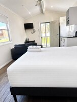 A modern hotel room with a large bed, a flat-screen TV mounted on the wall, a small kitchenette, and a sliding glass door to the outside.