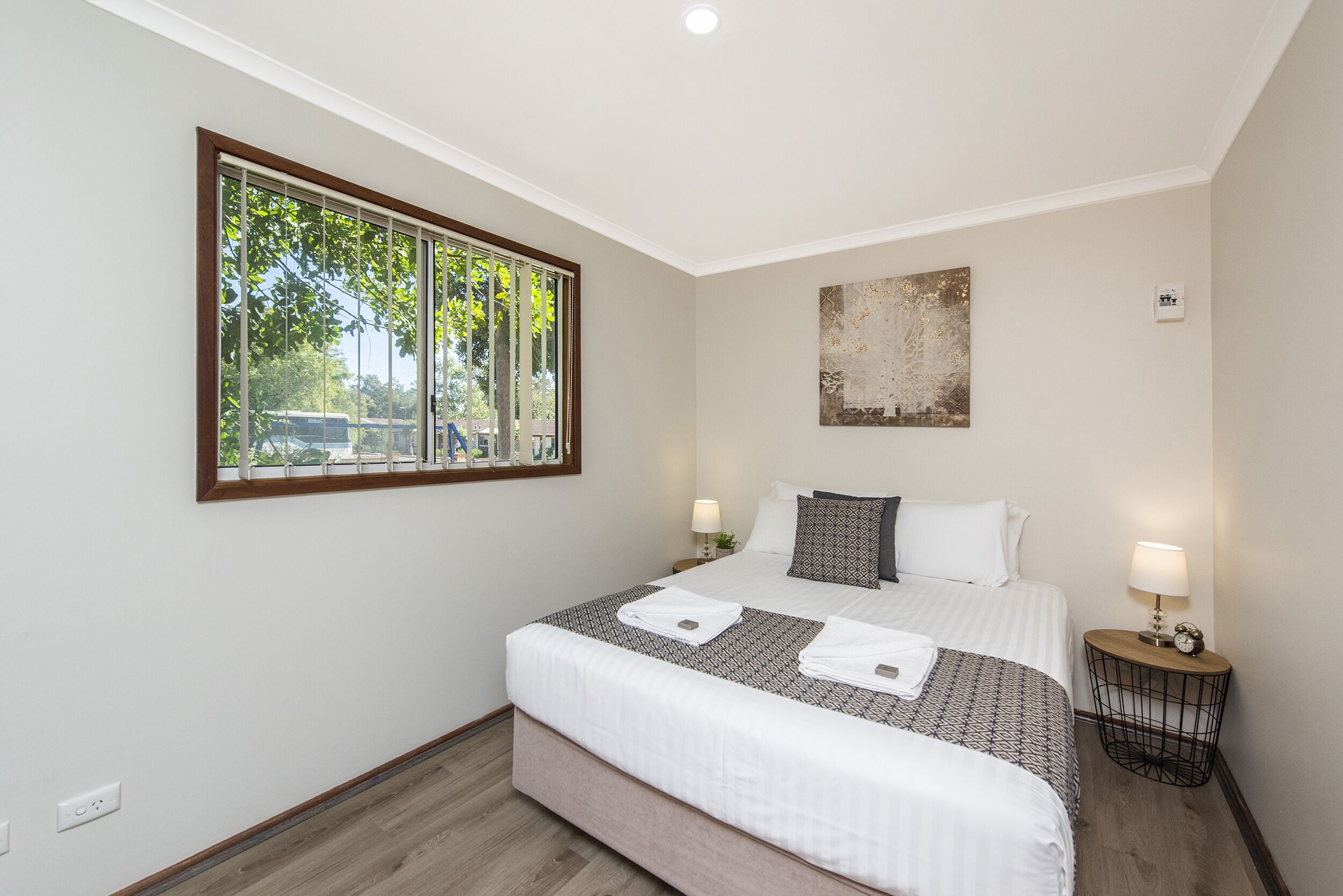 A neatly arranged bedroom with a large window, a bed with white linens, two bedside tables with lamps, and a wall-mounted artwork.