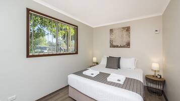 A neatly arranged bedroom with a large window, a bed with white linens, two bedside tables with lamps, and a wall-mounted artwork.
