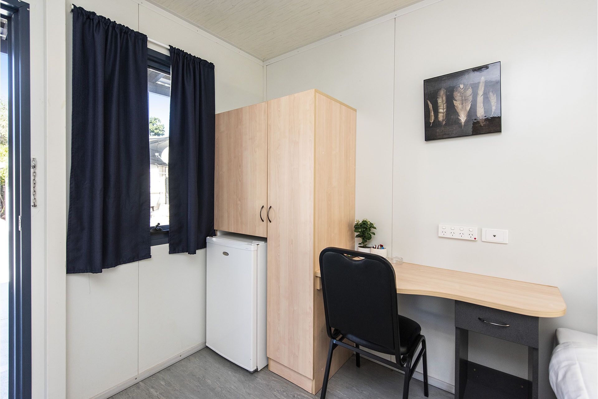A compact kitchenette with a refrigerator, a wooden wardrobe, a desk with a chair, and a small potted plant.