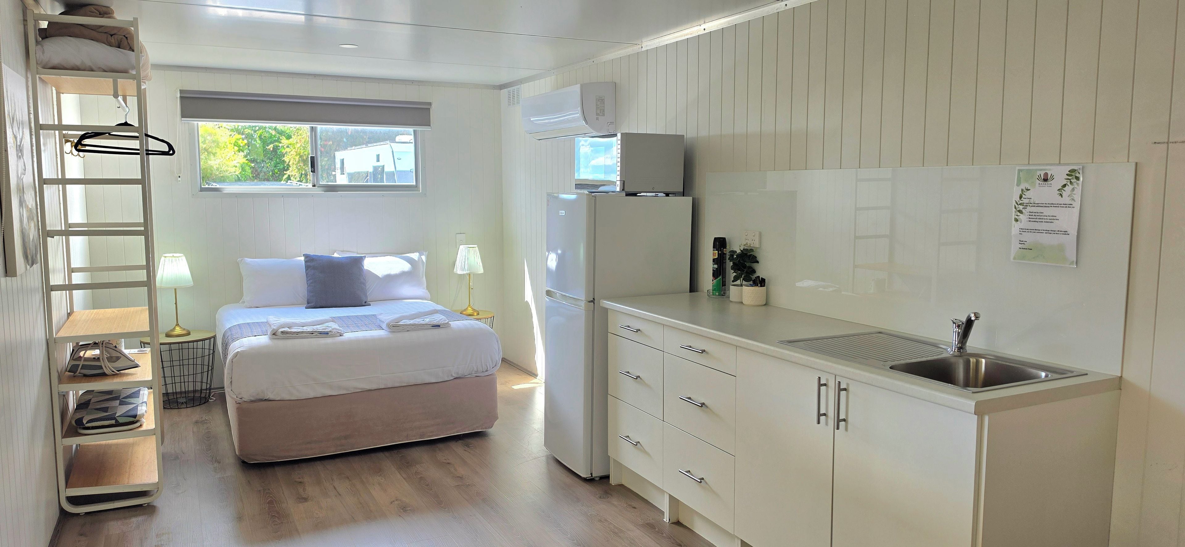A compact hotel room with a bed, bedside table, lamp, and a kitchenette area with a refrigerator and sink.