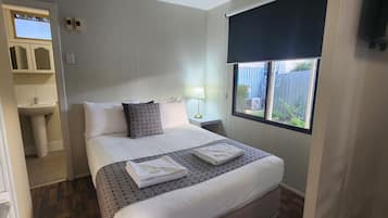 Quarto com cama, janela, mesa de cabeceira, abajur e banheiro visível por uma porta aberta.