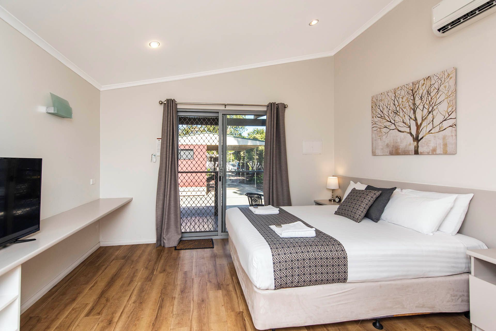 Habitación de hotel con cama, televisor, puerta con cortinas, un cuadro y vistas al exterior.