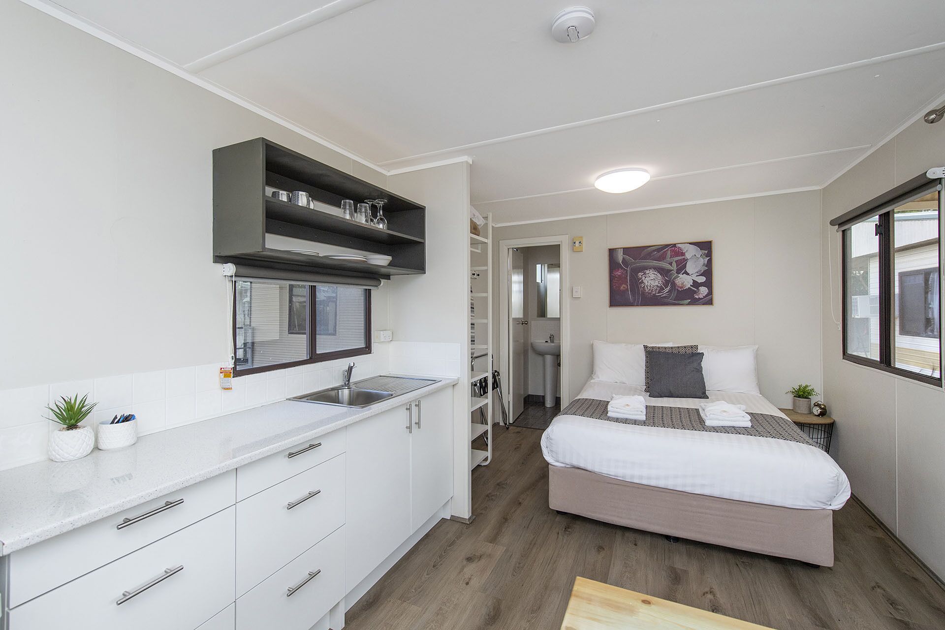 A compact hotel room with a kitchenette, a bed with white bedding, a small bedside table, and a window with a view.