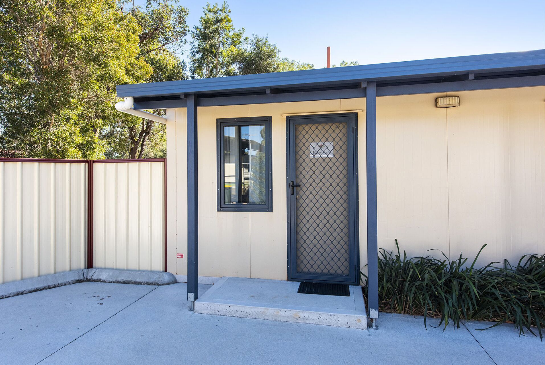 A small, single-story building with a metal roof and a beige exterior. It features a single door with a metal mesh screen and a window above it.
