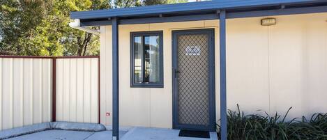 A small, single-story building with a metal roof and a beige exterior. It features a single door with a metal mesh screen and a window above it.