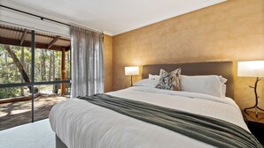 A spacious bedroom with a large bed, two bedside lamps, and a view of a forest through a sliding glass door. - Forest Rise (Yelverton)