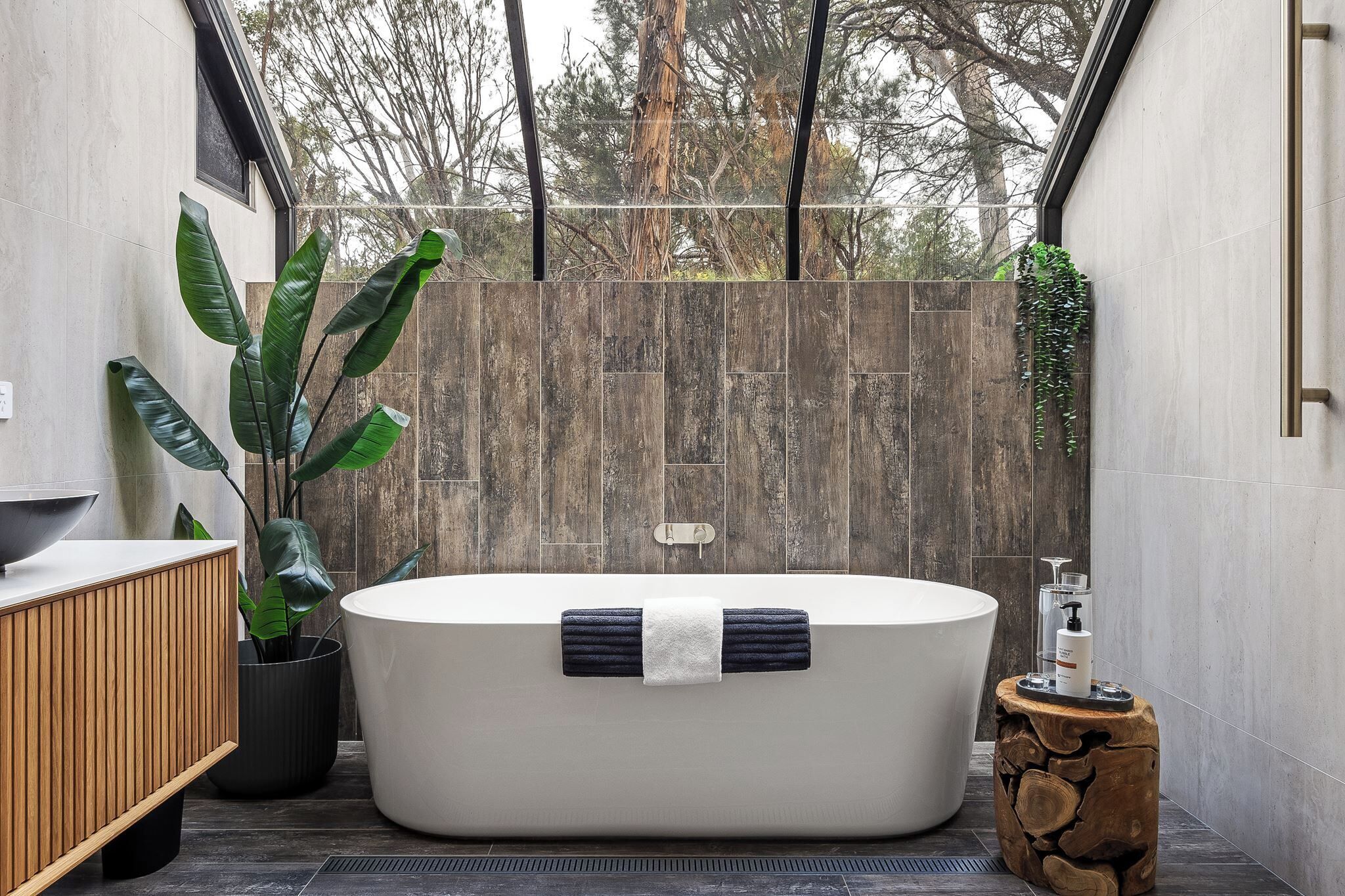 A modern bathroom with a freestanding bathtub, a wooden vanity, a potted plant, and a stone wall.