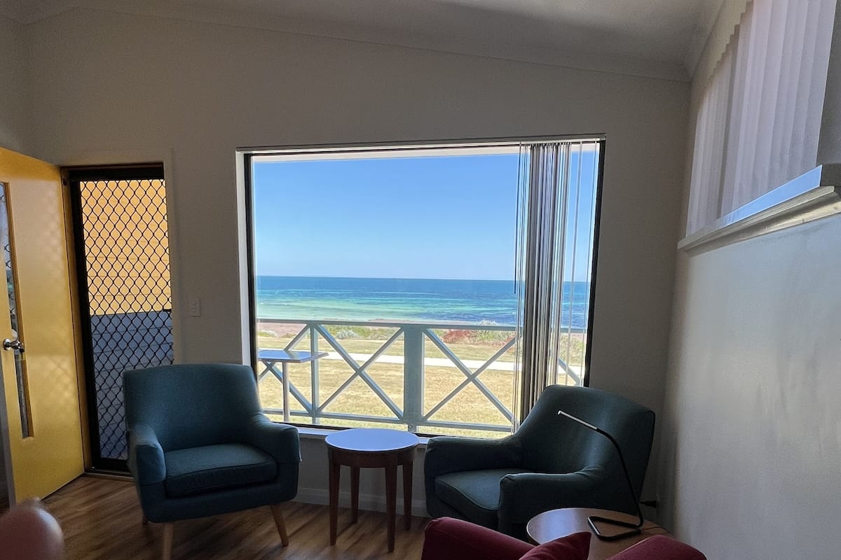 Ein Zimmer mit Holzboden, zwei Sesseln, einem roten Sofa und einem kleinen Tisch. Ein großes Fenster bietet Blick auf Strand und Meer.