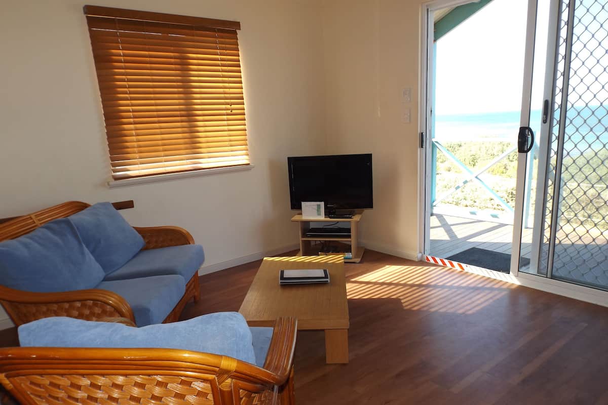 Ein Wohnzimmer mit einer Couch, einem Fernseher auf einem Schrank und Blick auf den Strand durch eine Glastür.