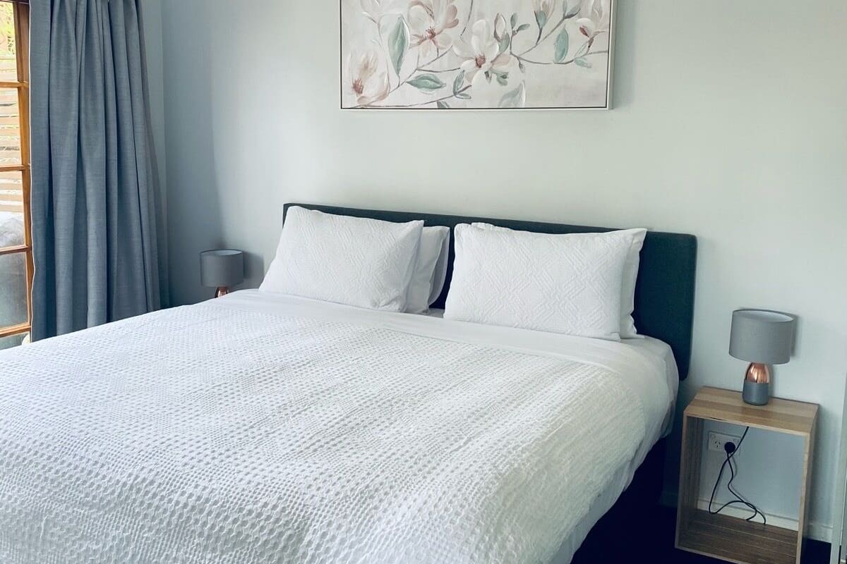 Une chambre à coucher avec un grand lit, deux tables de chevet, une lampe fixée au mur et une œuvre d’art à motifs floraux.