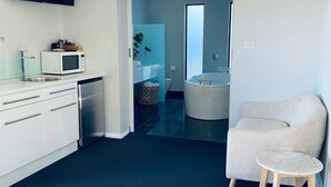Habitación de hotel moderna con cama, un pequeño espacio de descanso, kitchenette y baño visible a través de una puerta abierta.