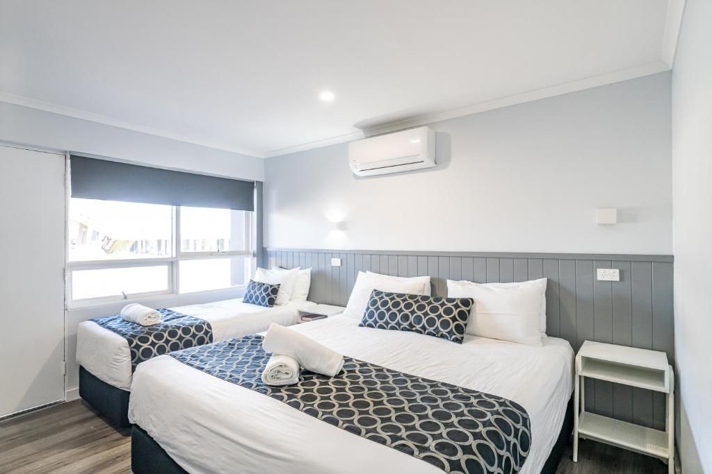A hotel room with two beds, a window, an air conditioning unit, and a small white nightstand.
