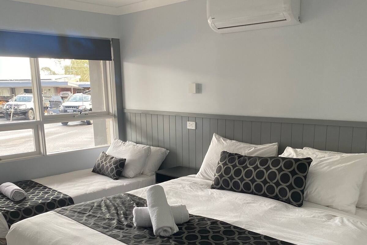 A hotel room with two beds, a window with a view of parked cars, a wall-mounted air conditioner, and a bedside table.
