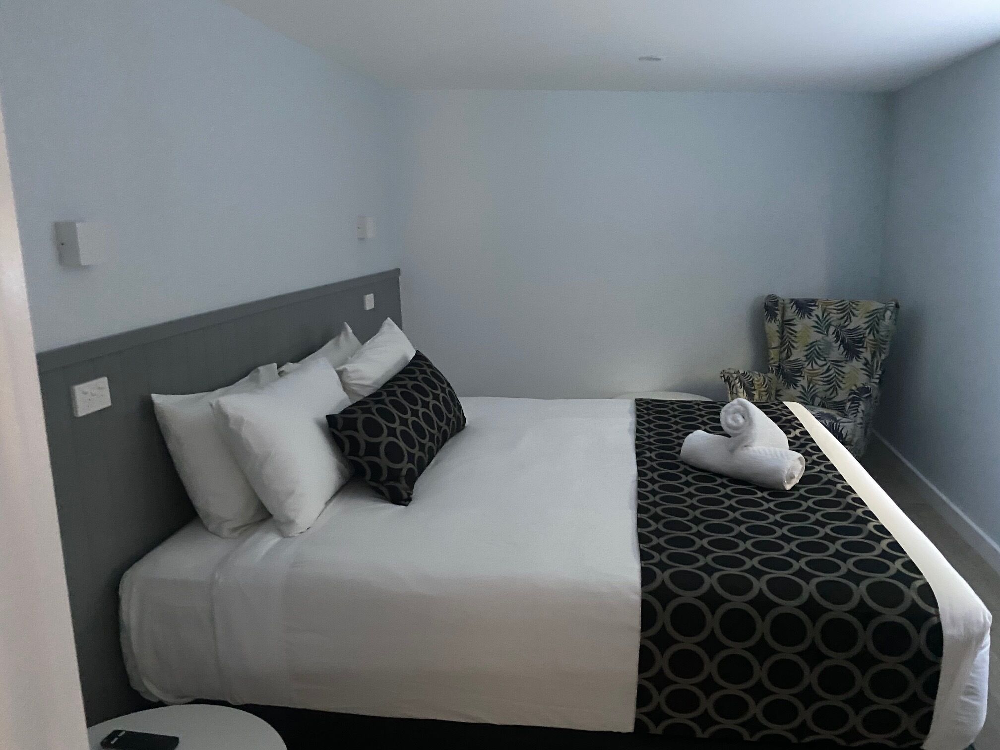 A hotel room with a bed, pillows, a patterned blanket, a chair, and a small table with a phone.