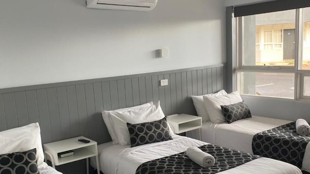A hotel room with two single beds, a small bedside table with a lamp, and a large window providing a view of the street. - Bega Motel (Bega)