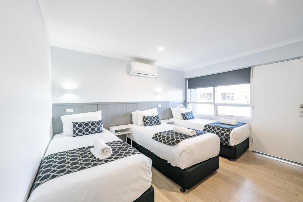 A hotel room with three beds, a window with blinds, an air conditioning unit, and a wooden floor.