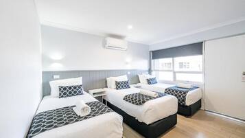 A hotel room with three beds, a window with blinds, an air conditioning unit, and a wooden floor.