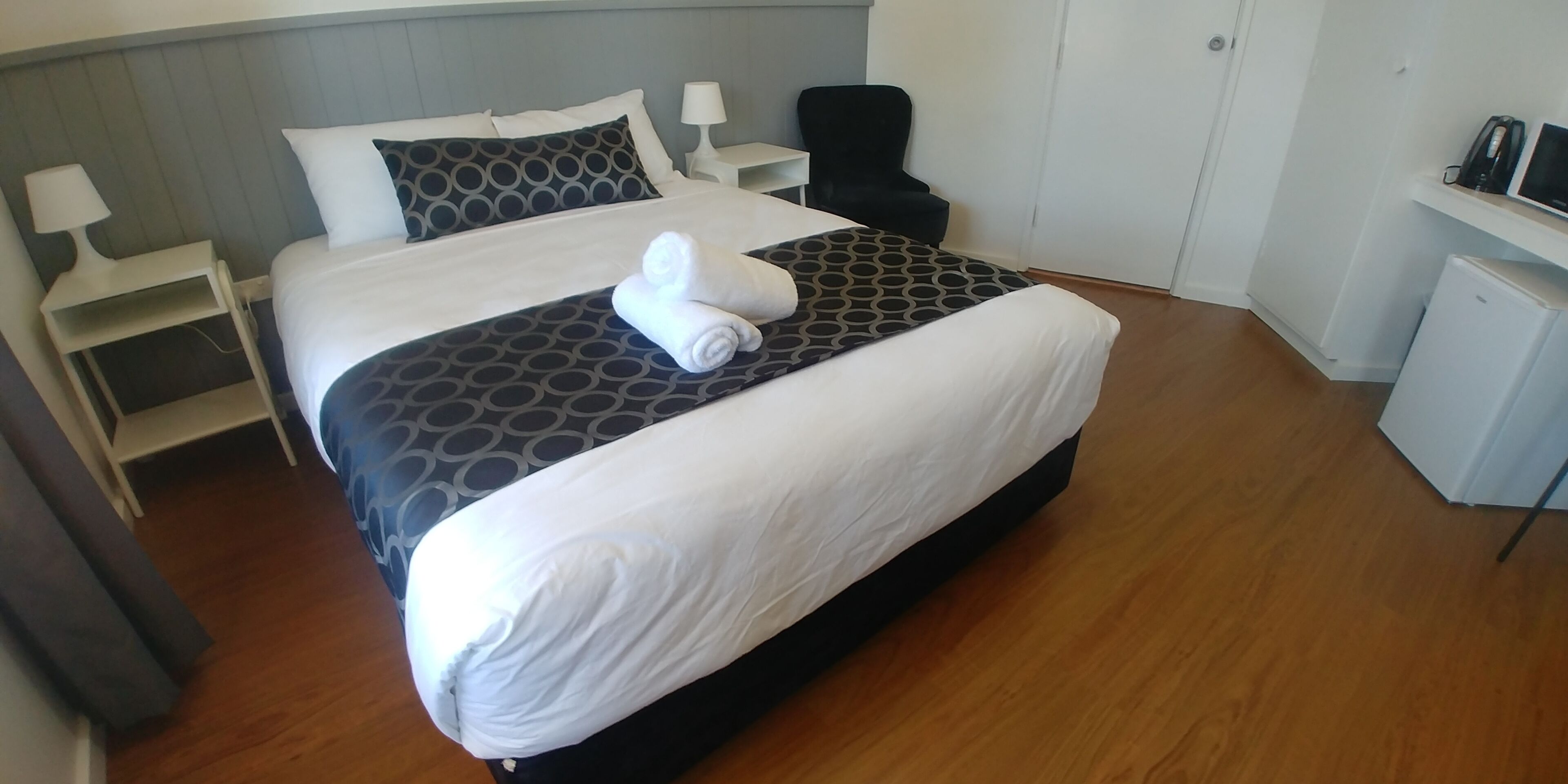 A neatly made bed with a patterned bedspread, a chair, a desk with a lamp, and a small refrigerator.