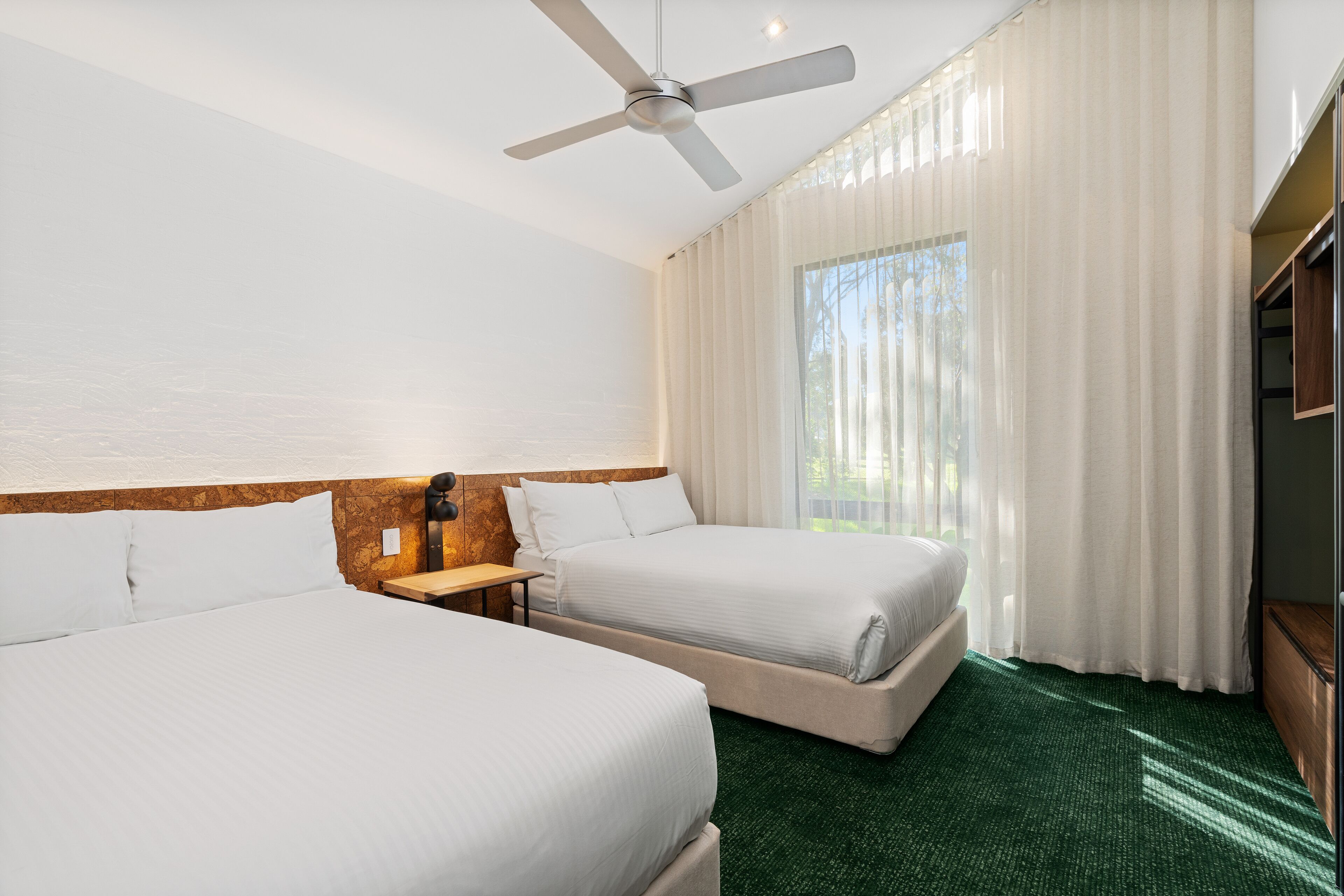A hotel room with two beds, a ceiling fan, a large window with sheer curtains, and a green carpet.