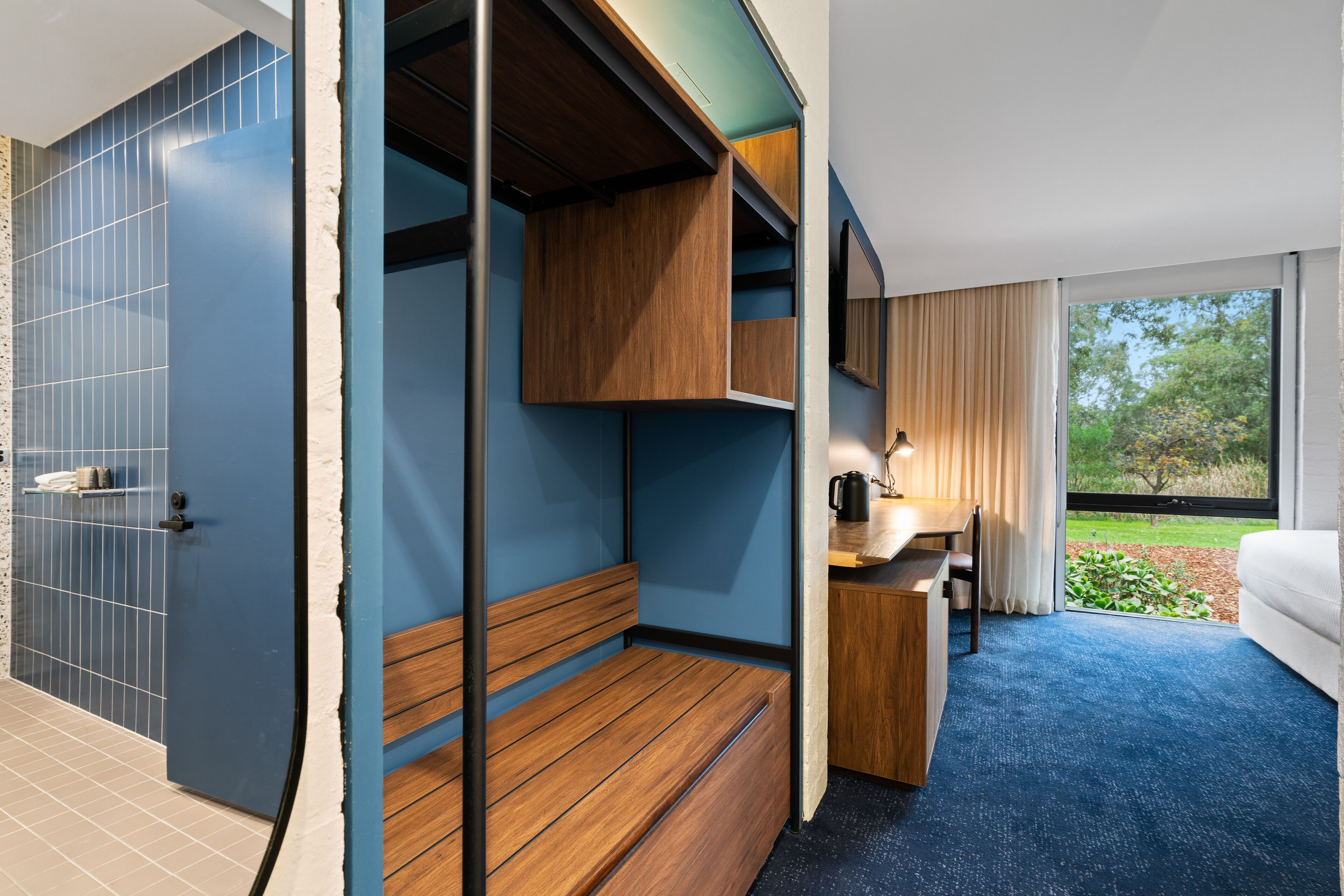 A compact hotel room with a built-in desk, a small seating area, and a view of the outdoors.