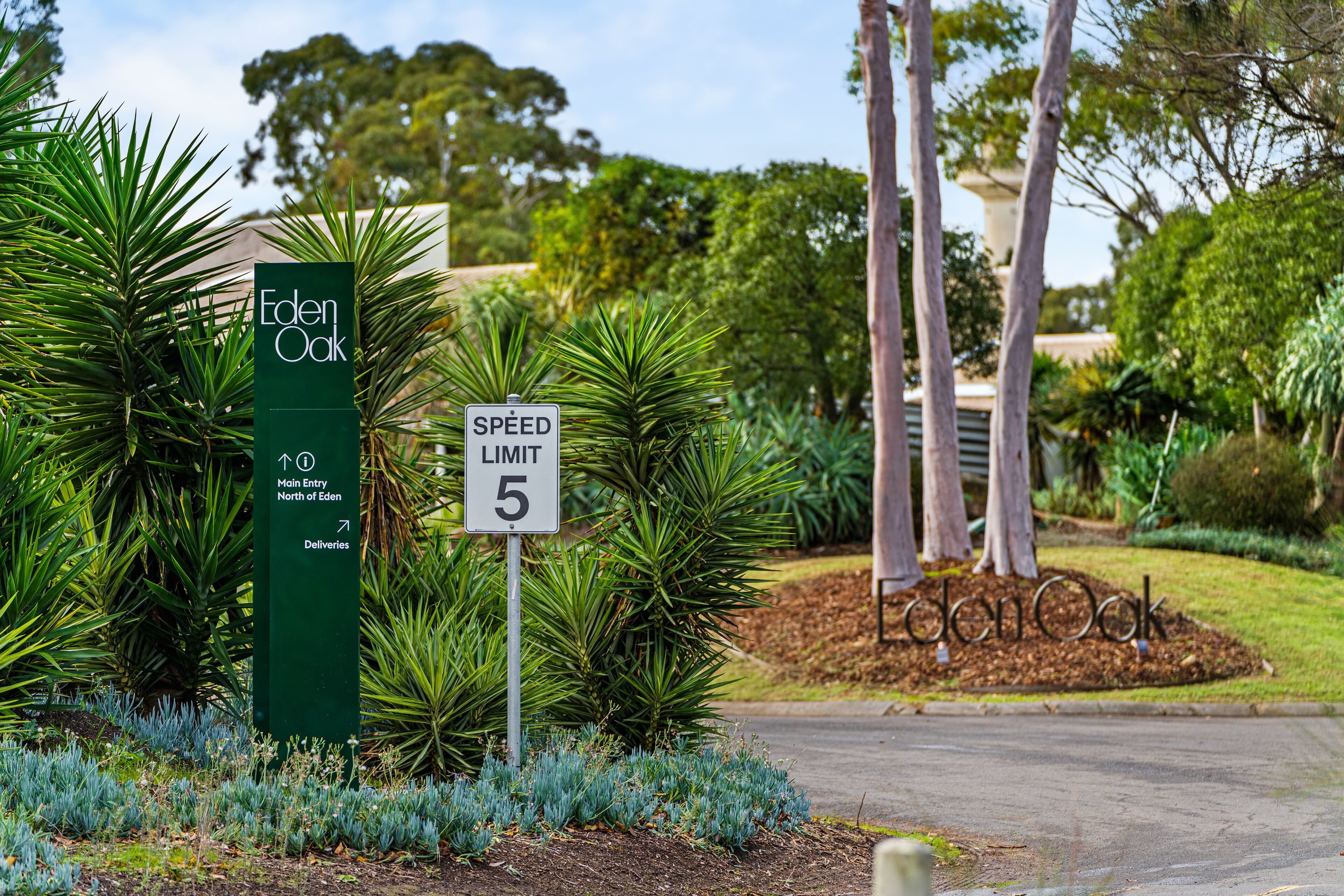 Una señal de límite de velocidad, un cartel verde con el texto 'Eden Oak' y una zona ajardinada con árboles y arbustos.