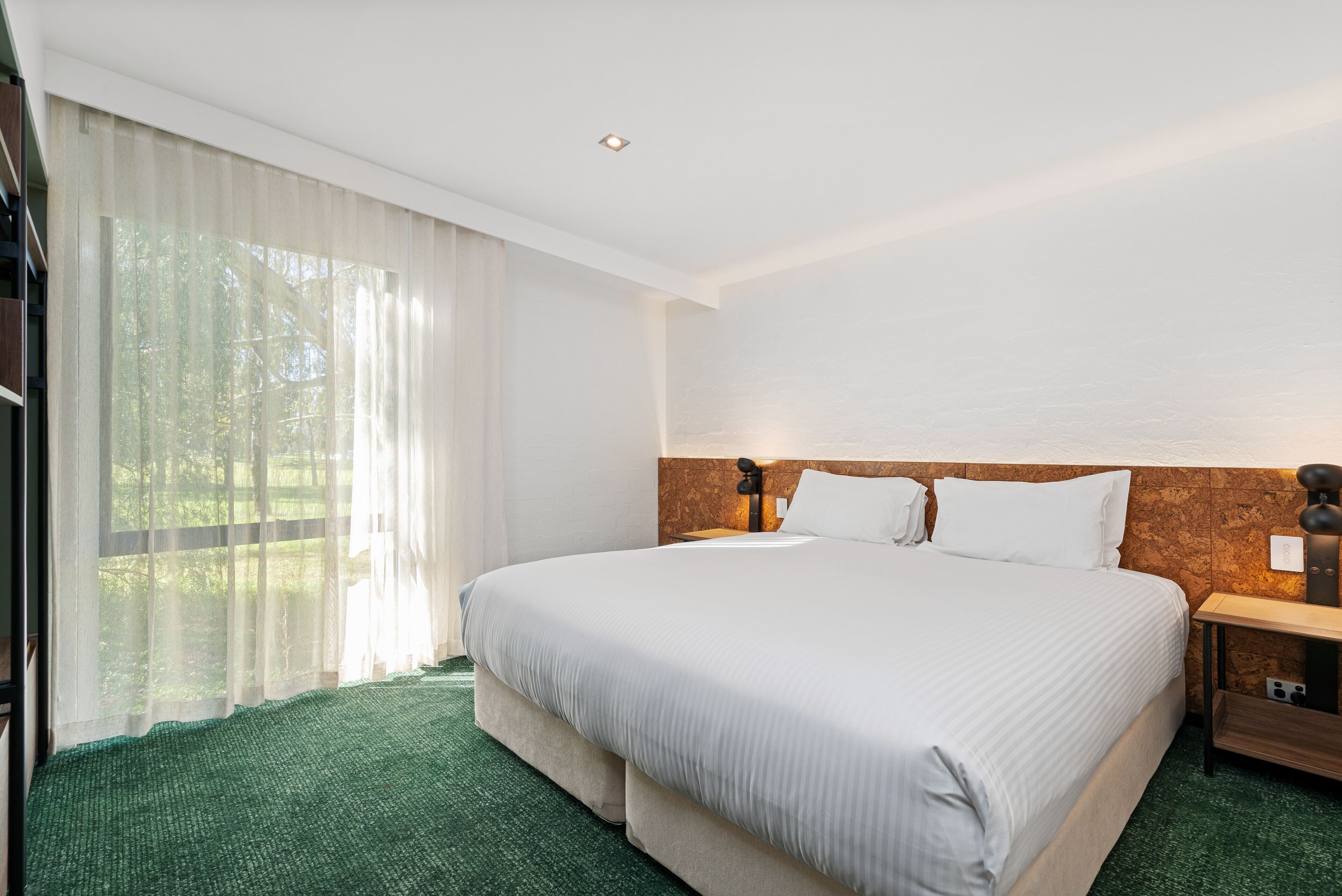 A hotel room with a large bed, a wooden headboard, a nightstand, and a window with sheer curtains.