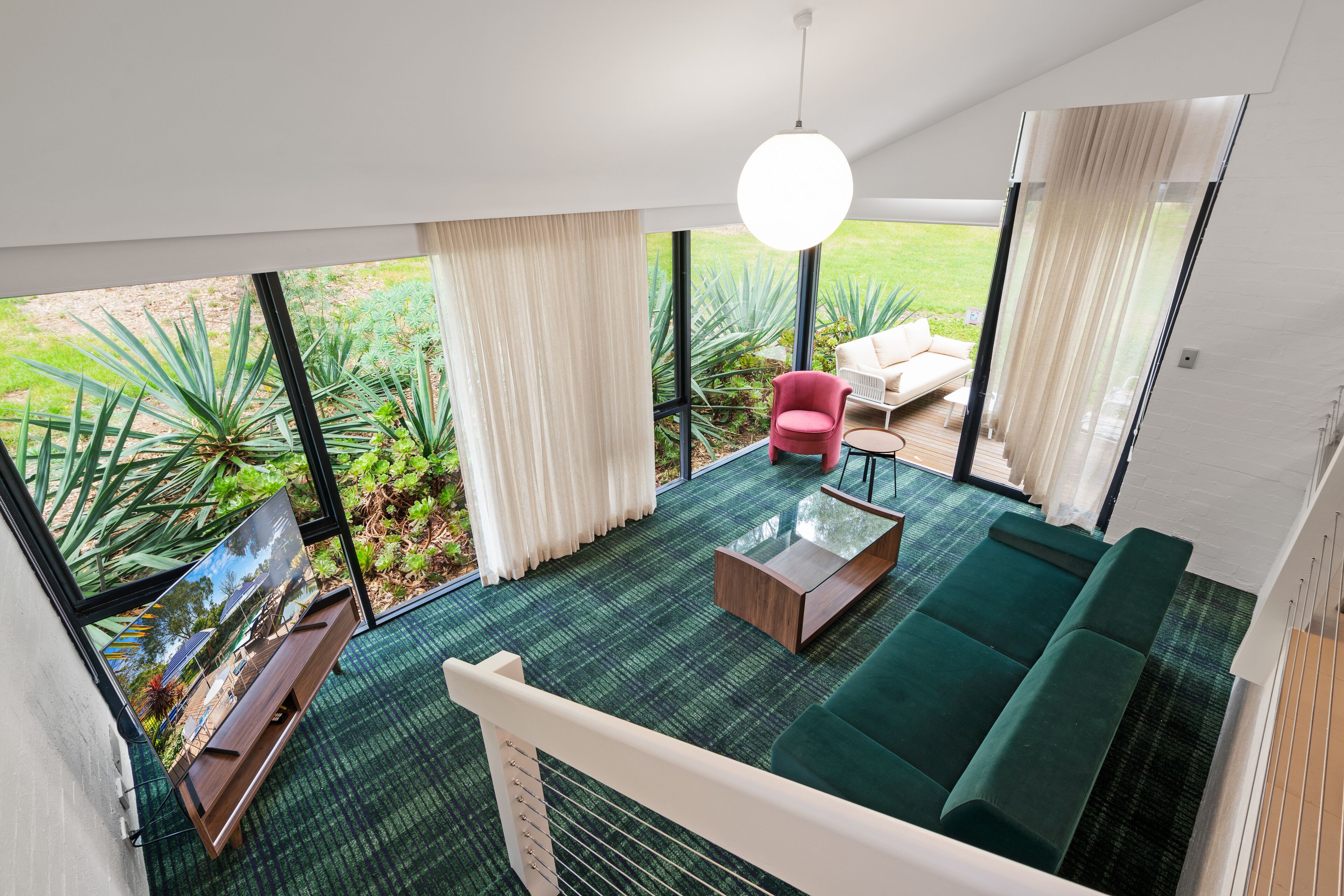 A modern living room with a green sofa, a red chair, and a wooden coffee table. Large windows offer a view of a garden.