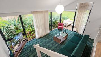 Un salon moderne avec un canapé vert, un fauteuil rouge et une table basse en bois. De grandes fenêtres offrent une vue sur un jardin.