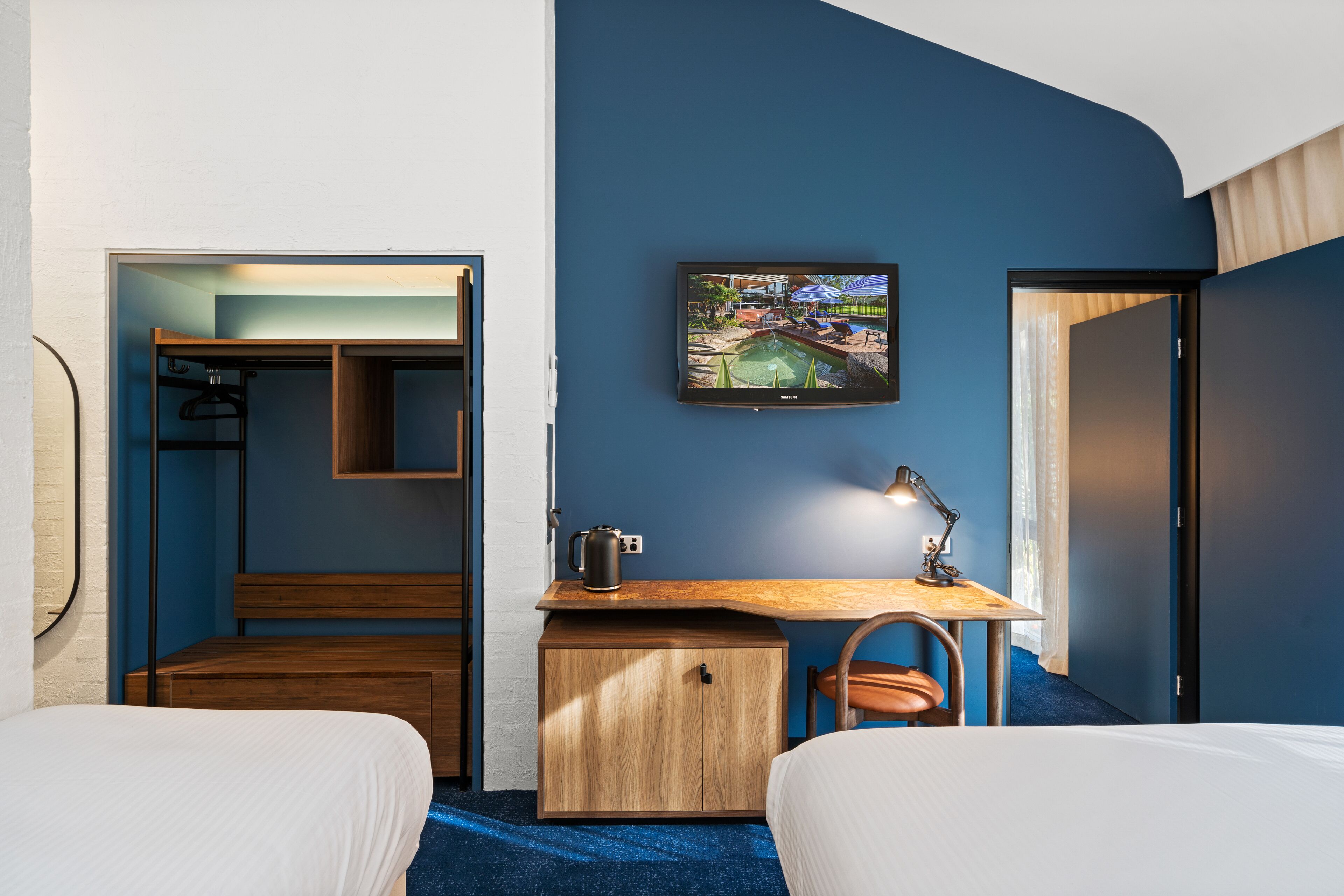 A hotel room with a bed, a desk with a lamp, a small cabinet, and a painting on the wall.