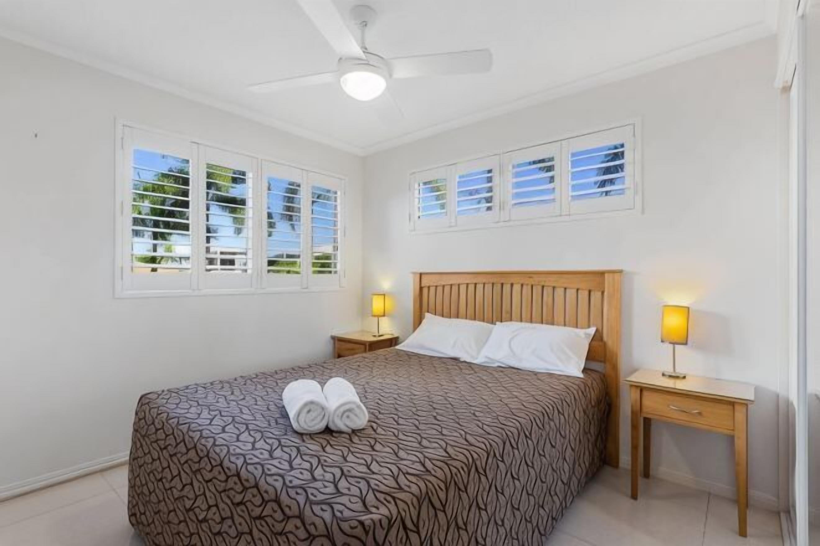 A bedroom with a bed, two bedside tables with lamps, a ceiling fan, and windows with blinds.