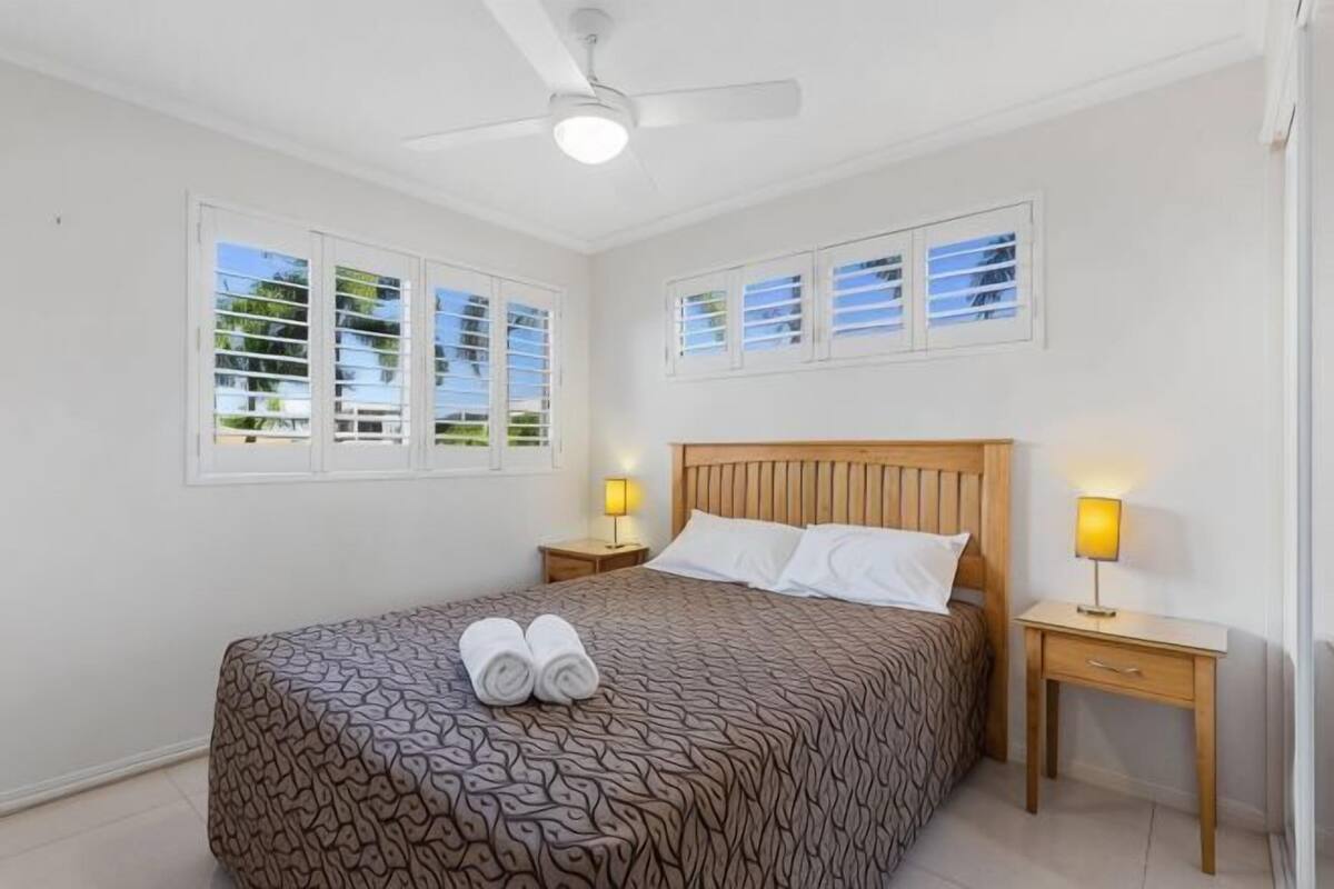 A bedroom with a bed, two bedside tables with lamps, a ceiling fan, and windows with blinds.