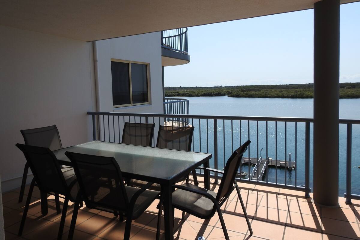 A balcony with a table and chairs, overlooking a body of water and a distant shoreline.