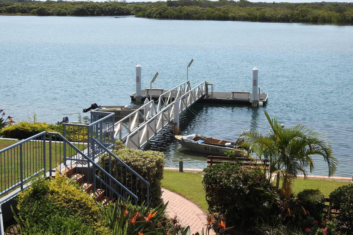 A dock extending into a calm lake with a bridge leading to a grassy area.