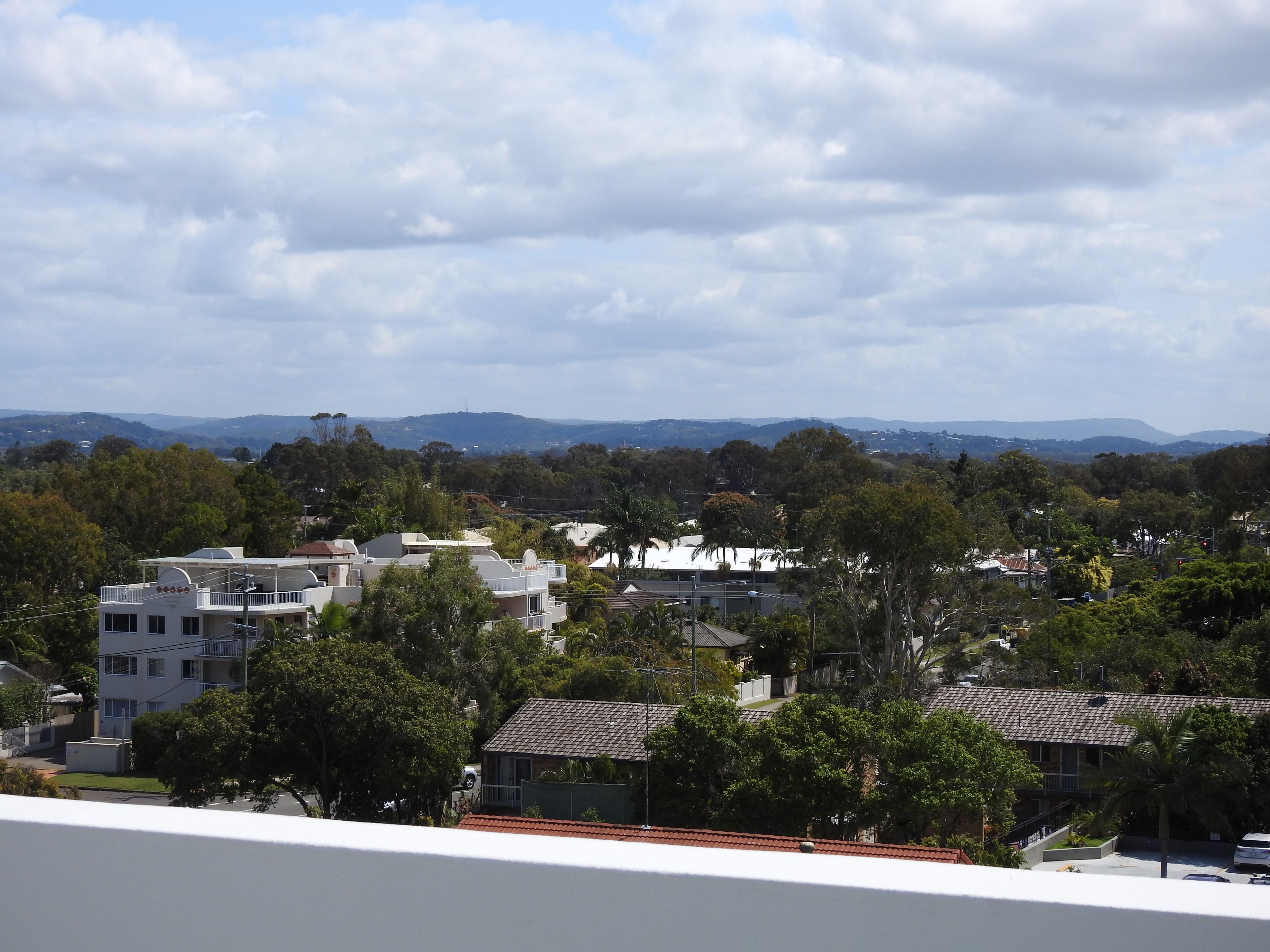 Un quartier résidentiel avec des maisons, des arbres et des montagnes lointaines, sous un ciel partiellement nuageux.