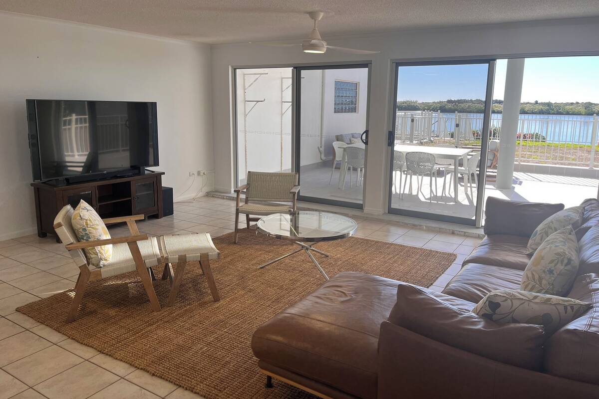 A living room with a TV, a sofa, a chair, and a glass table. There is a sliding glass door leading to an outdoor patio with a view of the water.