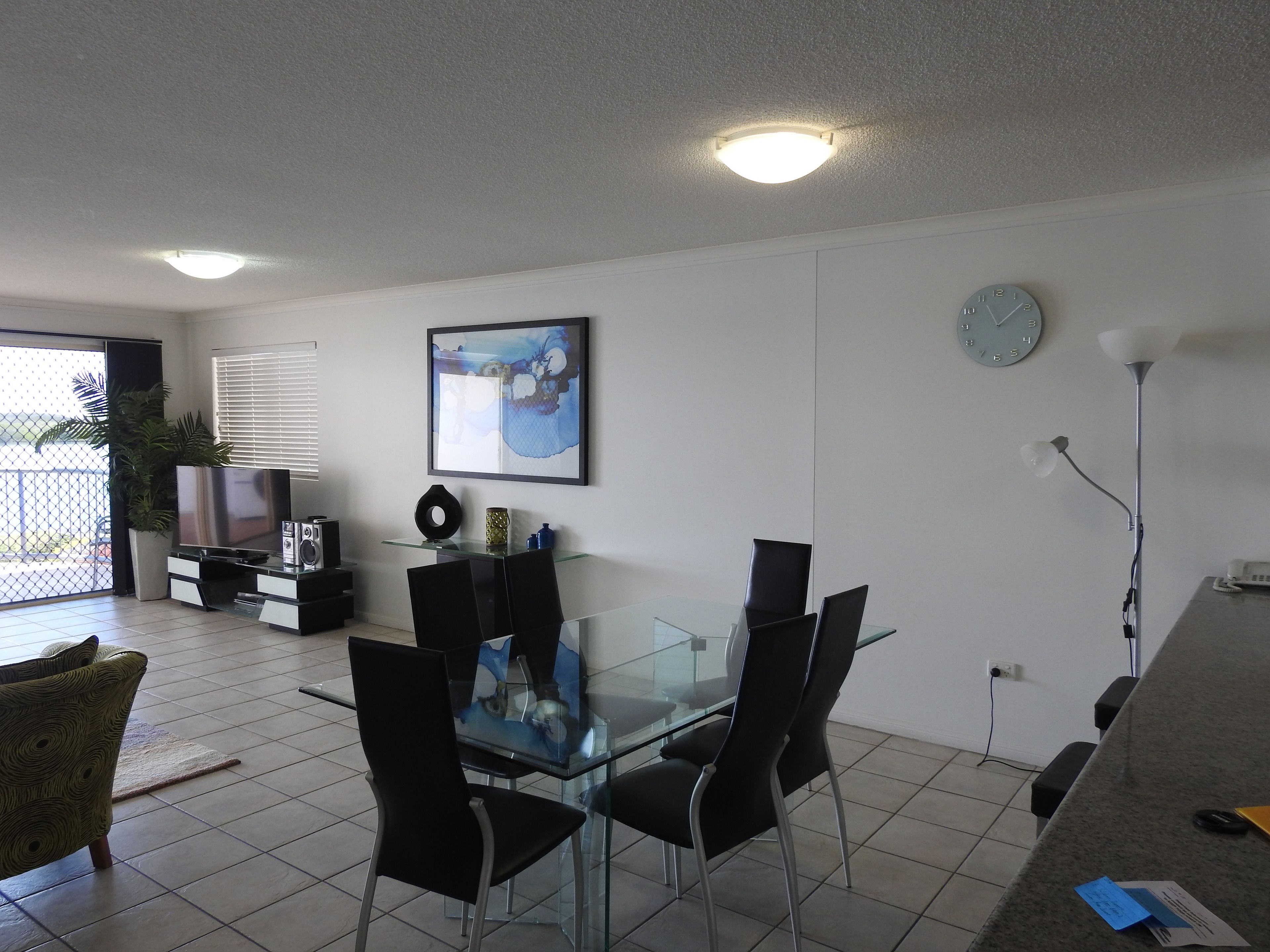 Un salon moderne avec une table à manger en verre, des chaises noires, un téléviseur à écran plat, un tableau, une horloge et une plante en pot.