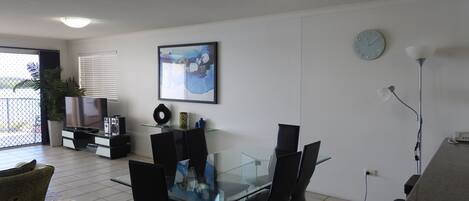 Un salon moderne avec une table à manger en verre, des chaises noires, un téléviseur à écran plat, un tableau, une horloge et une plante en pot.
