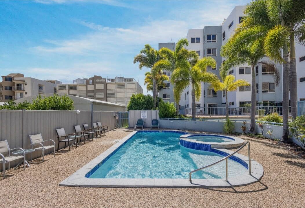 A swimming pool with lounge chairs and palm trees, surrounded by apartment buildings.