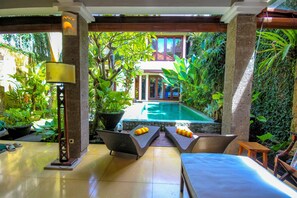 A pool area with lounge chairs and a table, surrounded by lush greenery and a view of a building in the background. - Umah Watu Villas (Seminyak)
