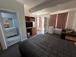 Un dormitorio con cama, televisor, refrigerador pequeño, sofá y baño con lavamanos y espejo.