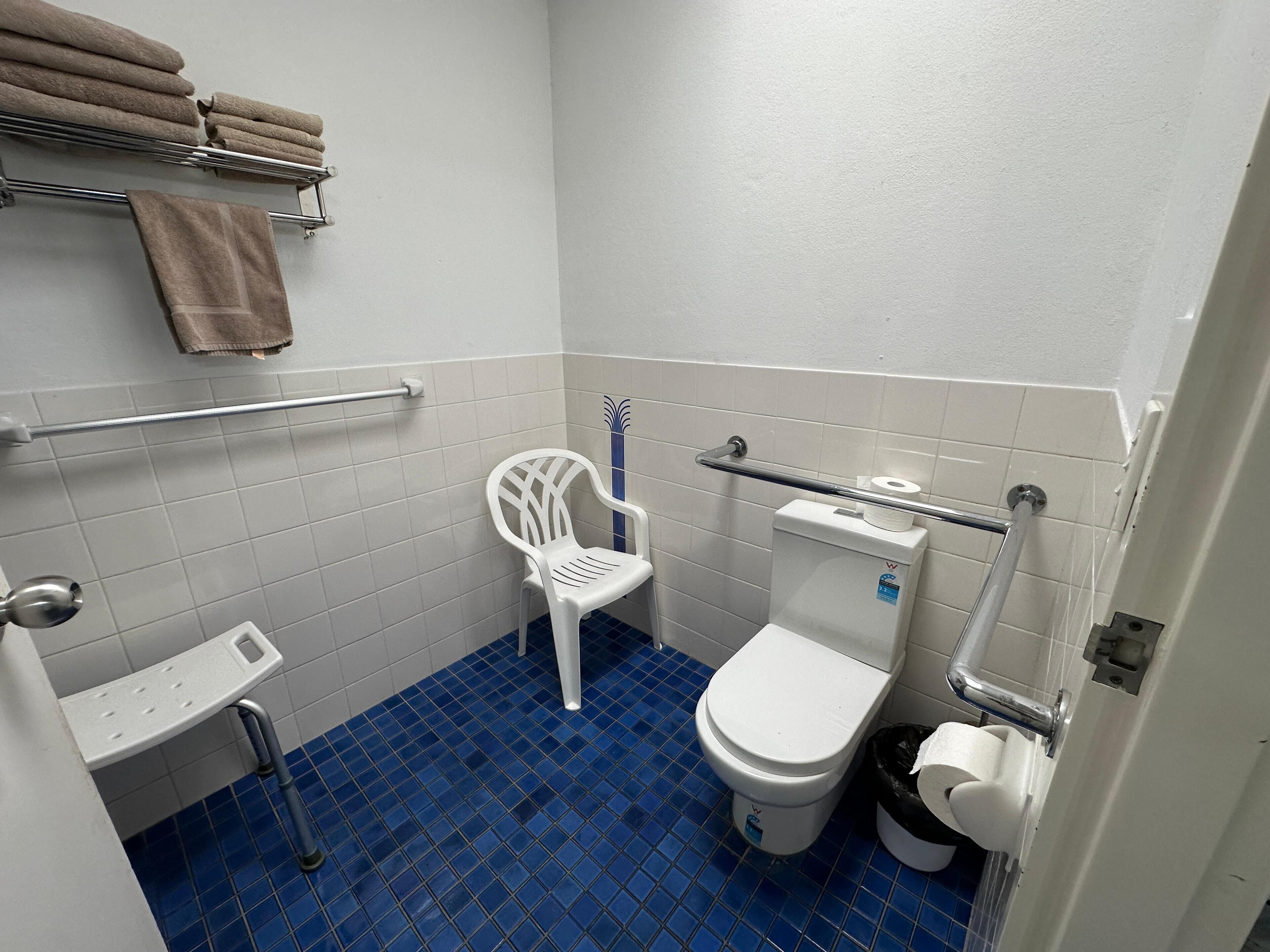 A small, clean bathroom with a toilet, a white plastic chair, a towel rack with towels, and a handrail.