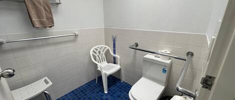 A small, clean bathroom with a toilet, a white plastic chair, a towel rack with towels, and a handrail.