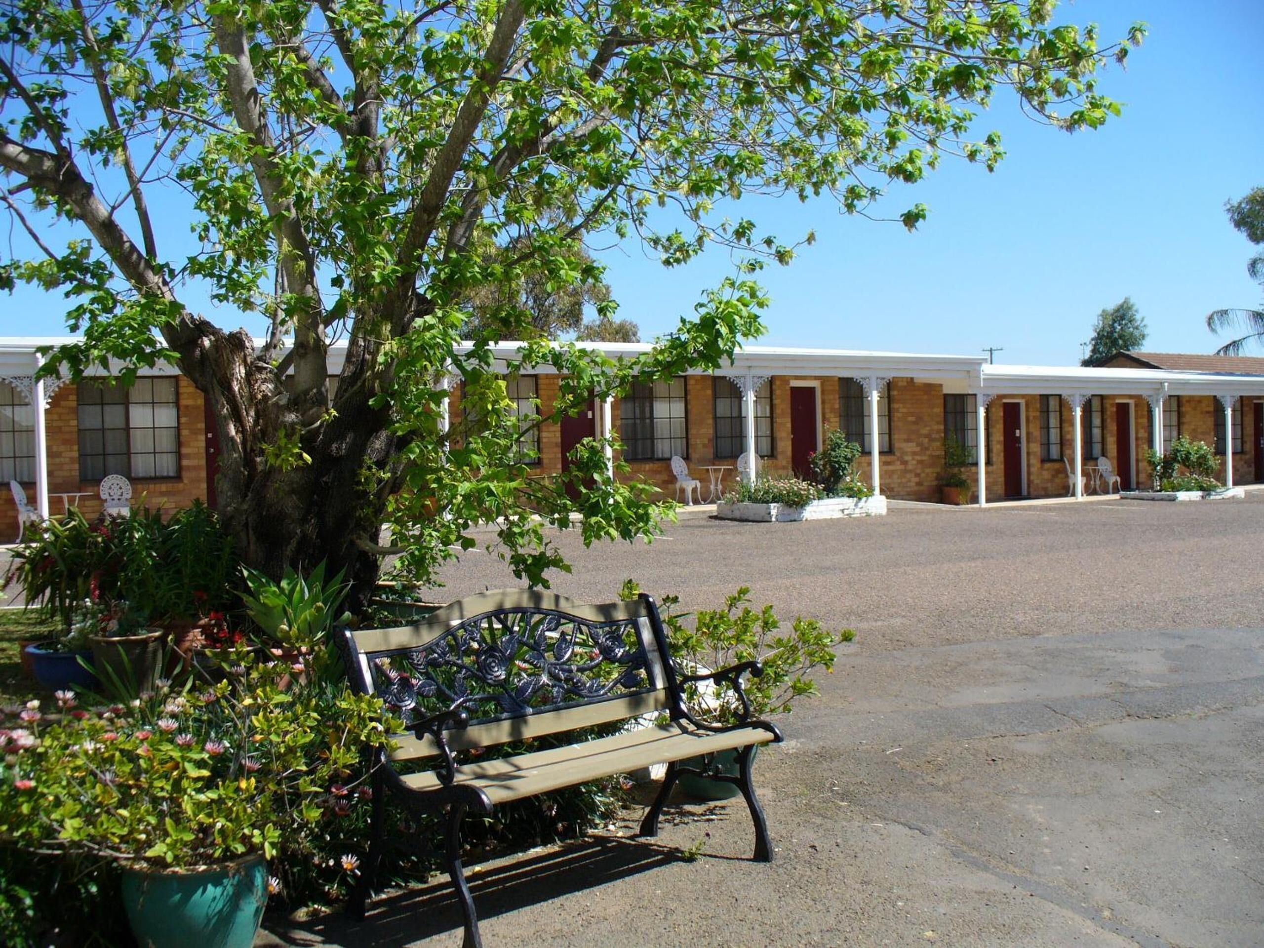A motel with a covered outdoor seating area, a large tree, and a bench.