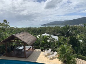 A poolside area with a thatched-roof structure, lounge chairs, and a view of the sea and hills.